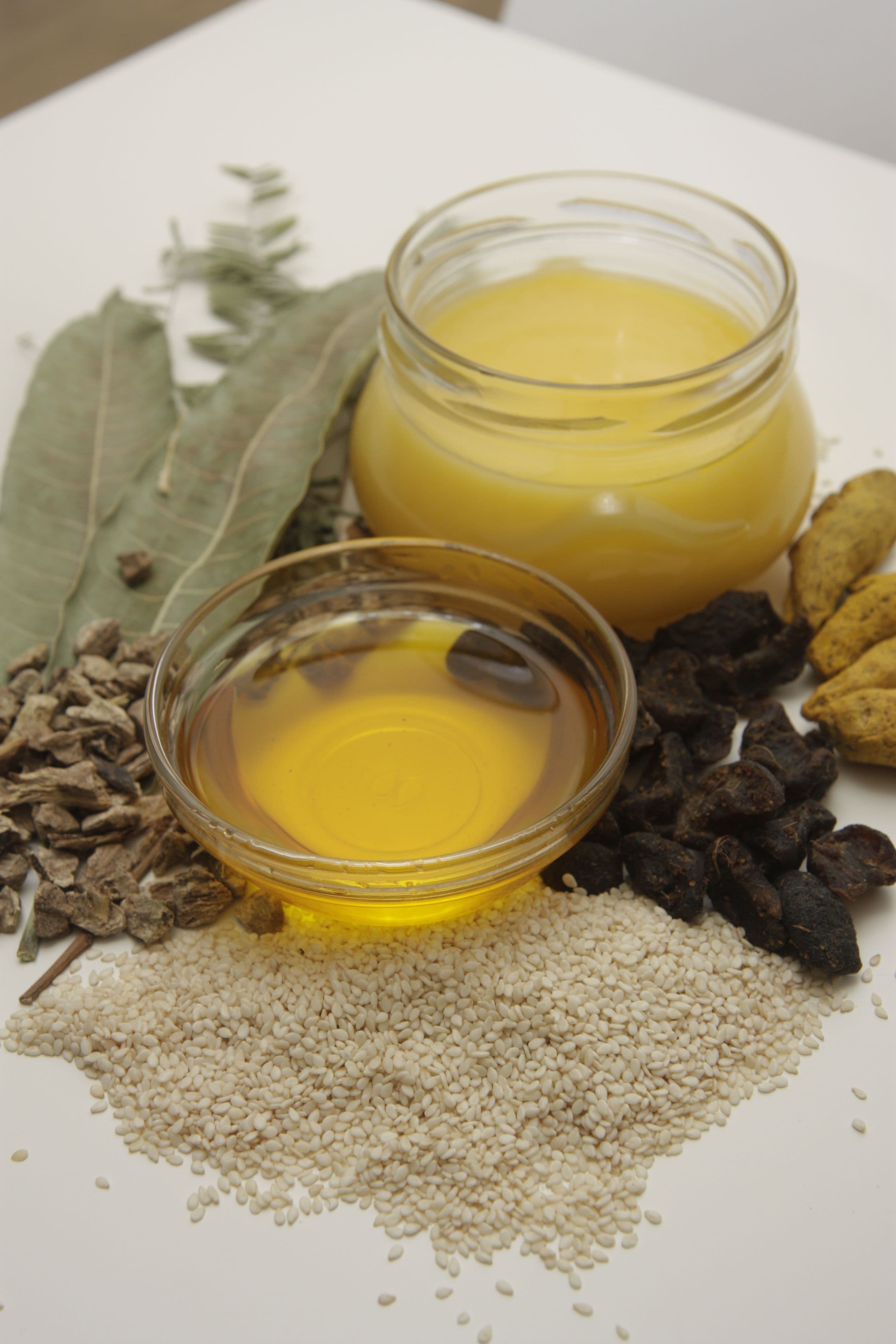 Ghee and sesame oil in glass bowls, set among sesame seeds, turmeric fingers and dried Ayurvedic herbs