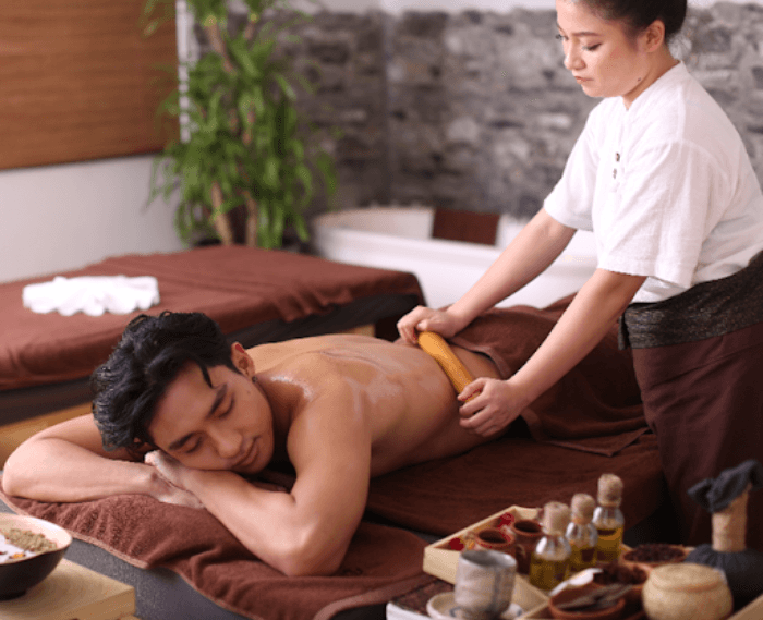A therapist working along an oiled back with a smooth wooden tool, oils and herbs on the tray alongside
