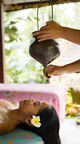 Warm oil running from a hanging copper vessel onto a guest's forehead, a frangipani flower in her hair