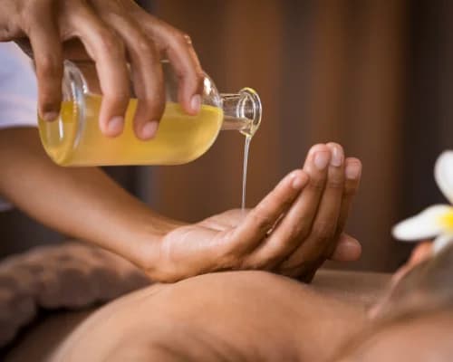 Warm herbal oil poured from a glass bottle into a therapist's cupped hand above a guest's back