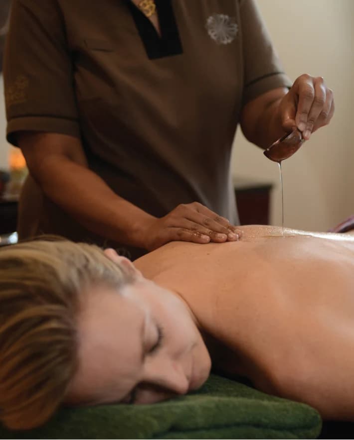 A therapist pouring a thread of warm oil along a guest's back during a full-body treatment