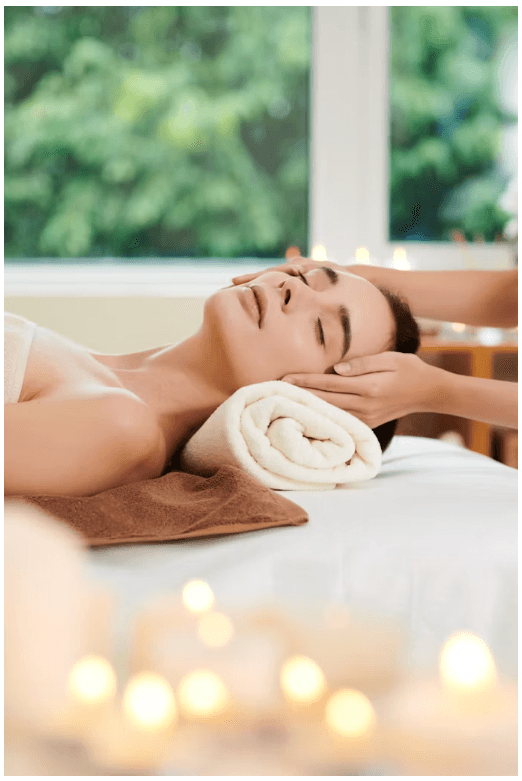 A guest resting on a treatment table with eyes closed while a therapist works along the jaw and temples, candles glowing in the foreground